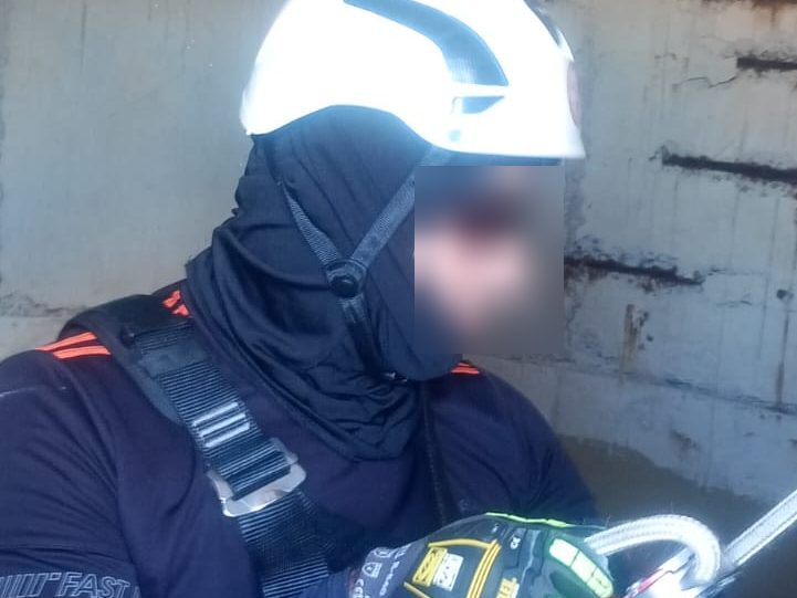 Profissional com capacete, balaclava e cinturão de segurança operando equipamento de acesso por corda dentro de reservatório de concreto, com o rosto desfocado por privacidade