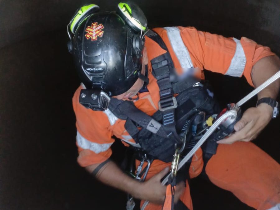 Profissional de uniforme laranja com capacete e cinturão de segurança conectado à corda, iniciando a descida pela abertura de um reservatório cilíndrico de concreto, com a identificação do uniforme desfocada
